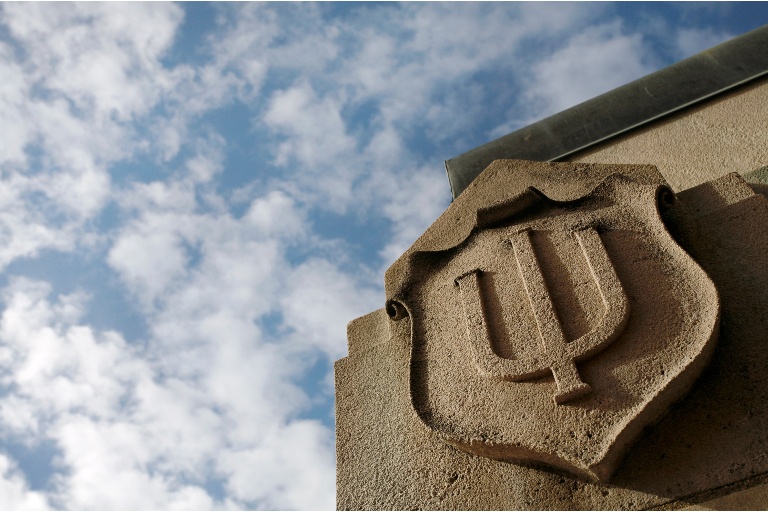 IU logo on a shield carved in limestone on the Bloomington campus. Bright blue skies in the background.